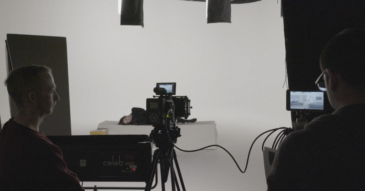 Behind the scenes photo taken from behind a video camera looking at an actress sleeping on a bed.