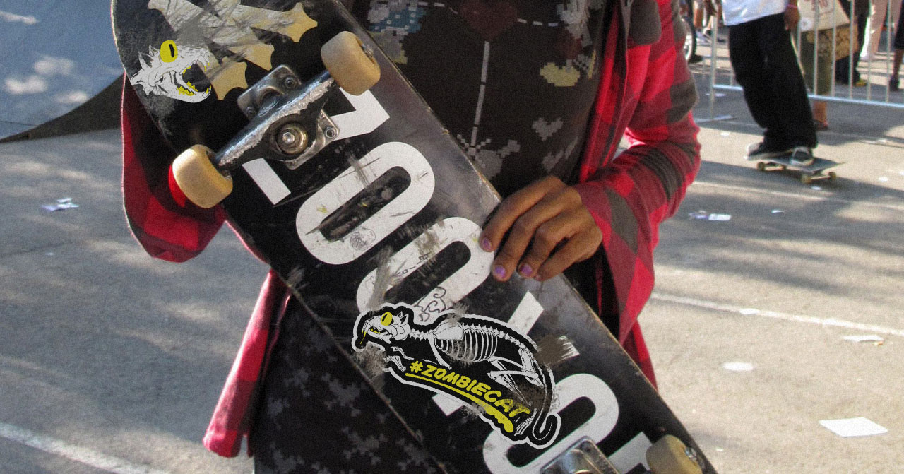 Young person holding a skateboard with #zombiecat stickers on it.