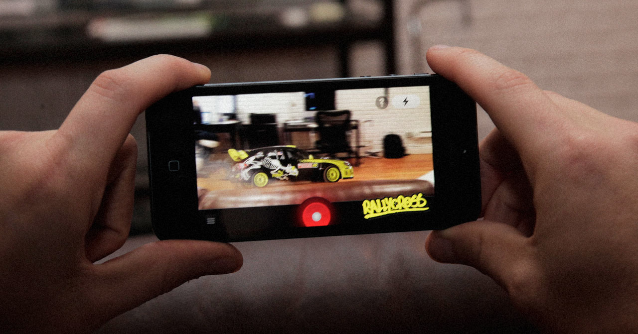 Close up of two hands holding a phone displaying a small car driving over a couch in augmented reality