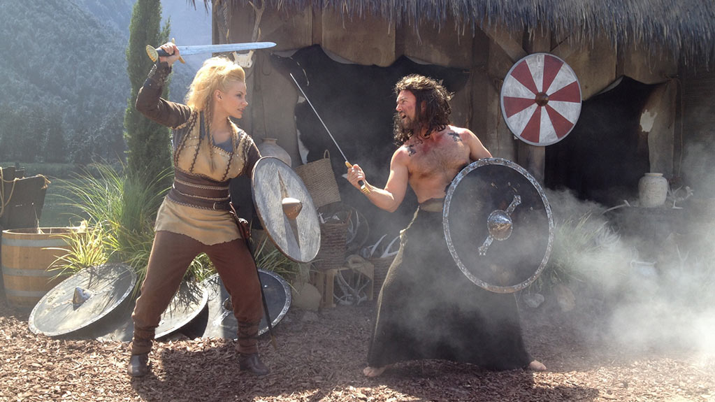 Two people dressed as vikings swordfight in front of a hut.